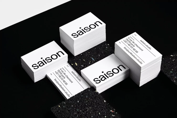 Stacks of minimalist business cards on a black surface, featuring the word 'saison' and contact information for saison.studio in Paris.