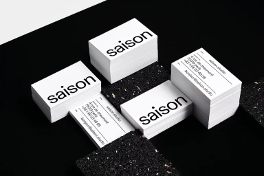 Stacks of white business cards with black typography displaying Saison branding arranged on a dark textured surface