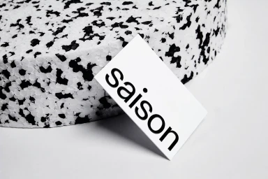 A white card with the black text Saison resting against a speckled black and white textured circular foam block on a white background