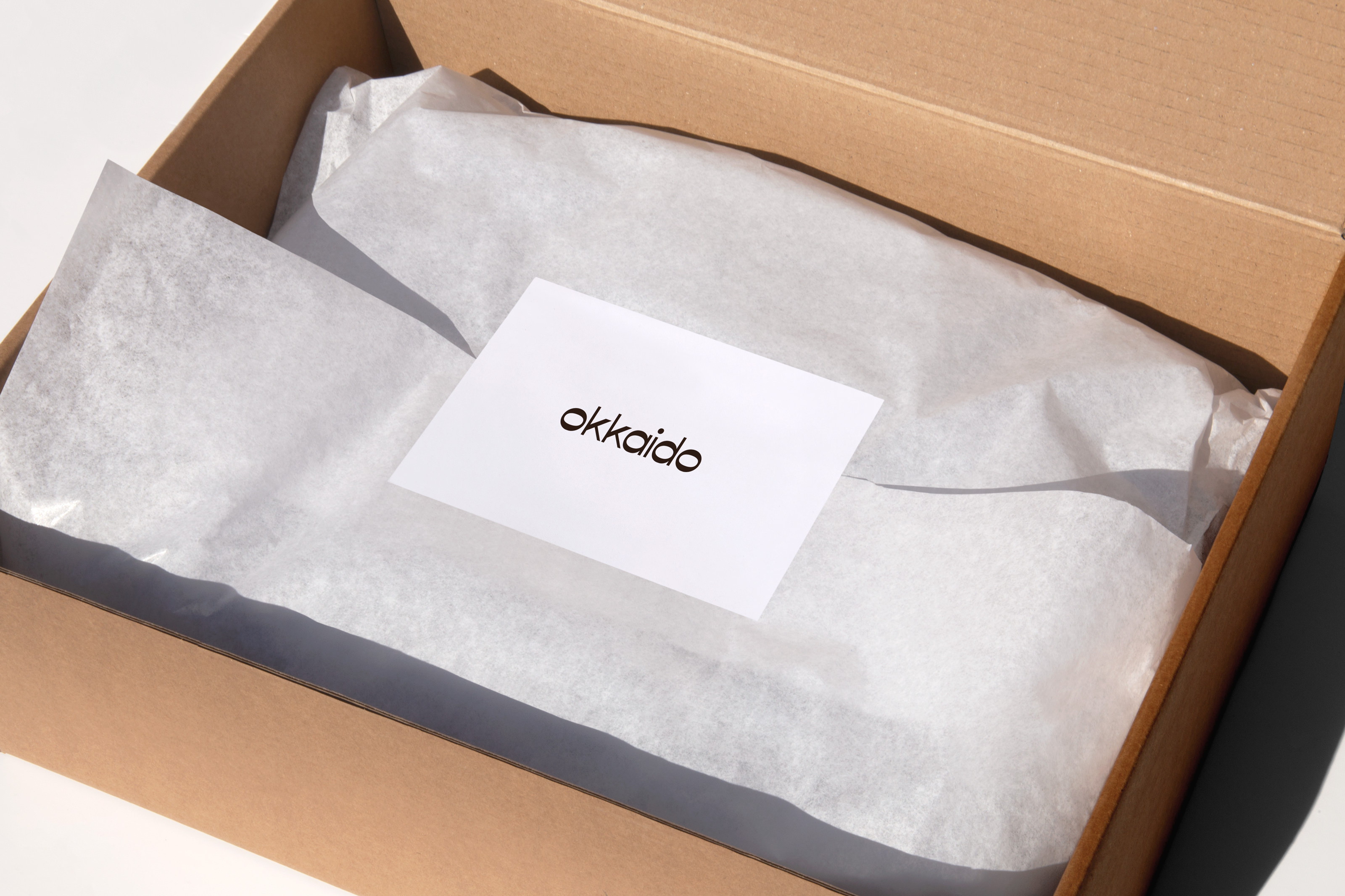 An open cardboard shipping box shows crumpled white tissue paper and a company card reading "okkaido" against a white background.