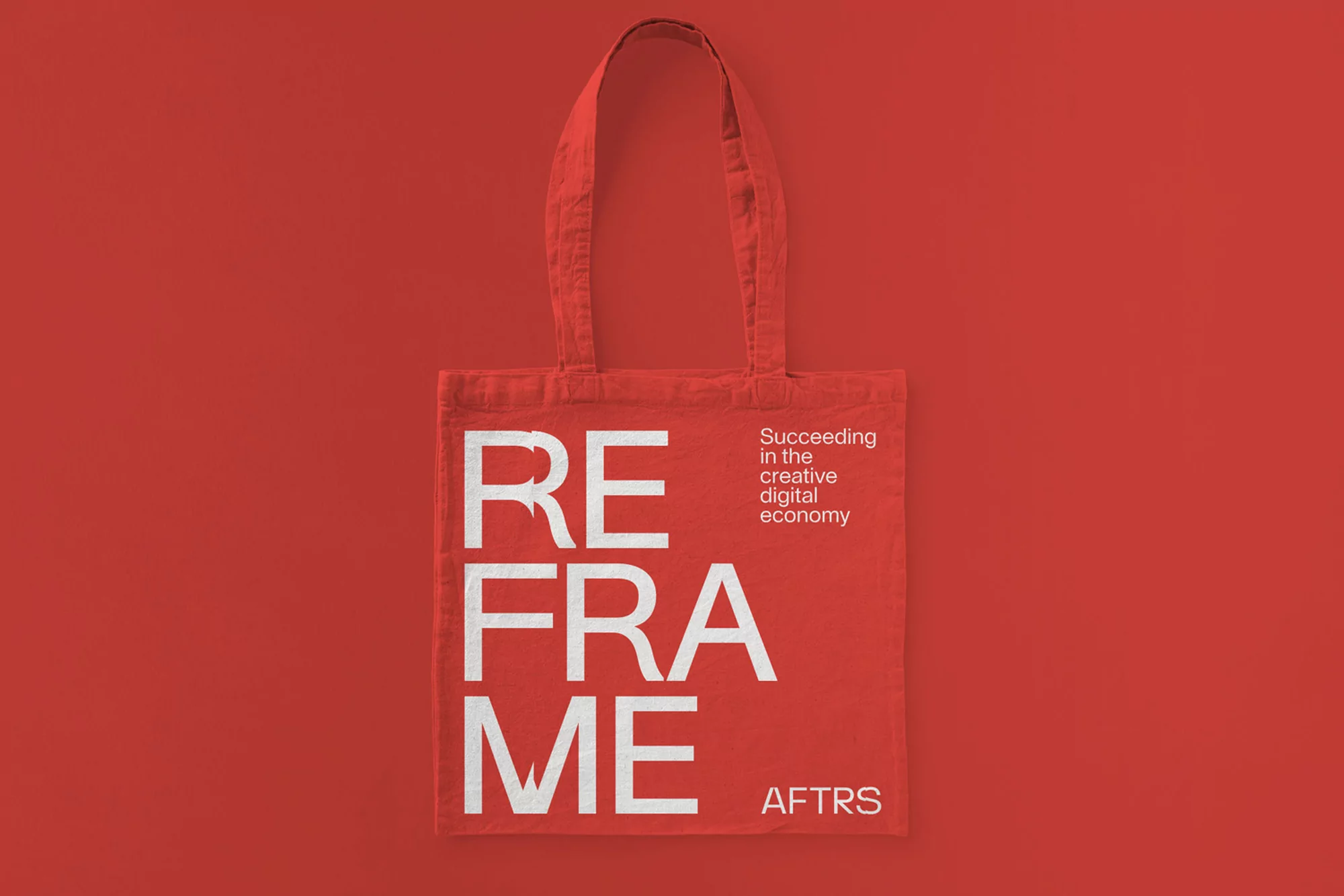 Red tote bag with white 'Reframe' text and 'Succeeding in the creative digital economy' text displayed against a solid red background.