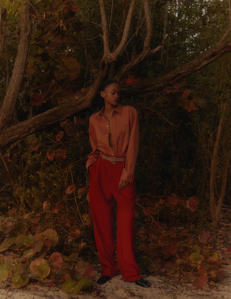 A young Black woman in a rust-colored shirt and red pants stands in a natural outdoor setting surrounded by foliage and bare branches.