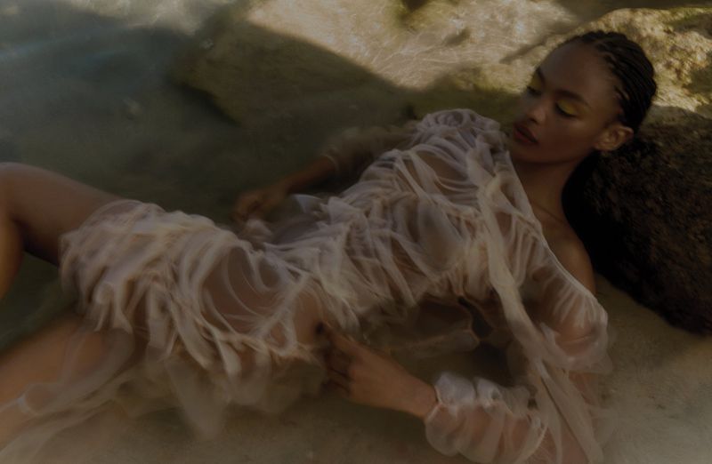 An adult woman with braided hair lies in shallow water, wearing a diaphanous white dress, rocks in the background, medium shot, eye-level.