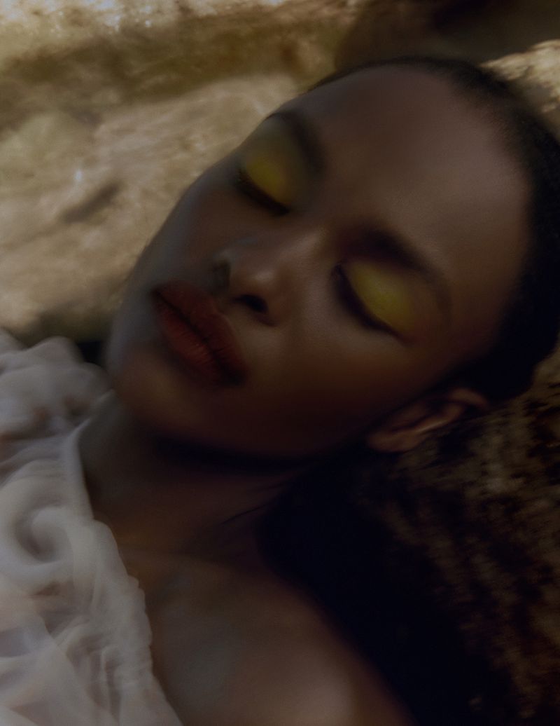 Closeup portrait of a serene woman with bold yellow eyeshadow and red lipstick lying down with a blurred beige background and white fabric.