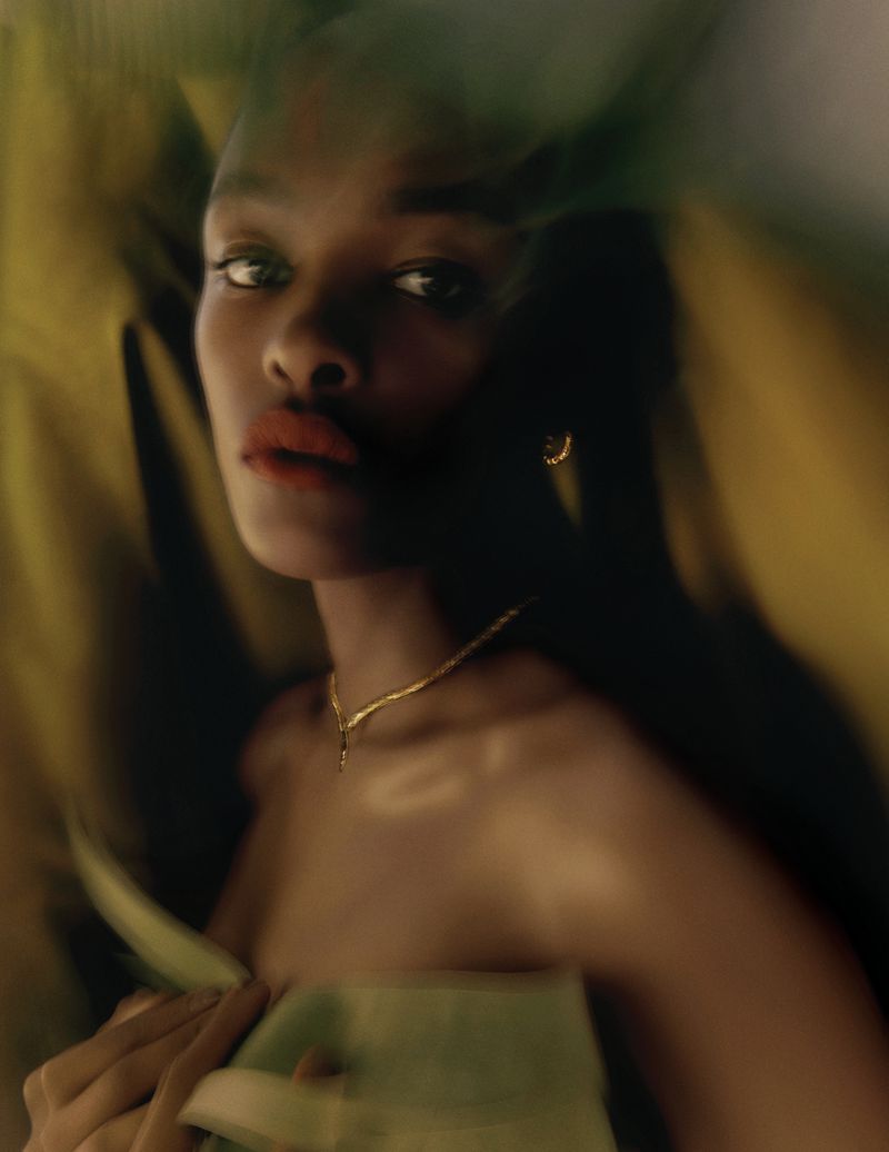 Portrait of an African American woman with red lipstick and golden jewelry, posing against a blurred green and yellow background.