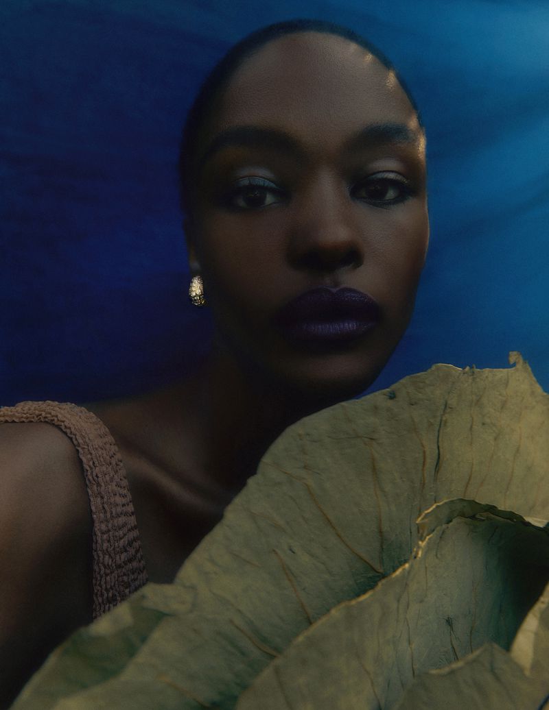 Close-up portrait of a dark-skinned woman with purple lipstick posing with a large green leaf against a blue backdrop.
