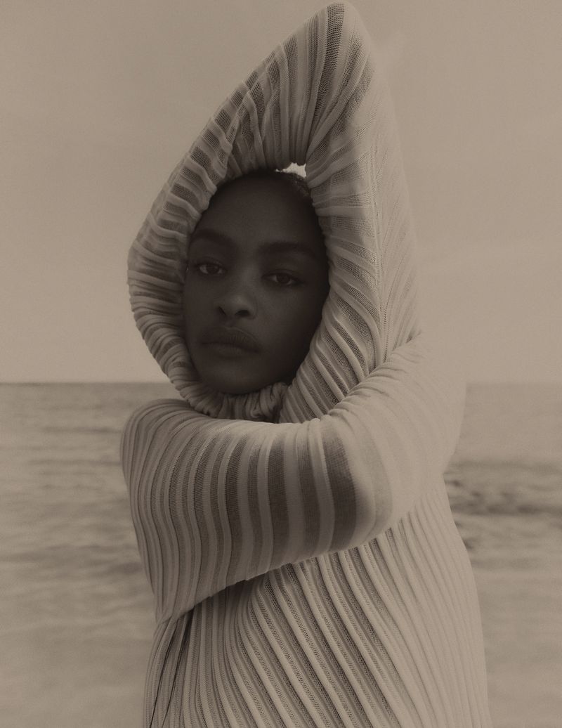 Monochromatic portrait of a woman in draped, textured fabric against a serene seascape, showcasing a blend of fashion and nature.