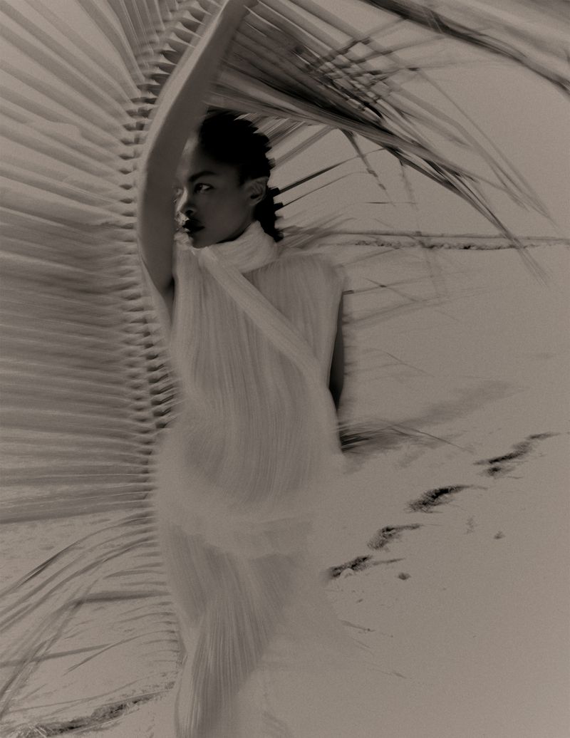 Woman in a light dress poses with a large palm frond on a sandy beach in a monochrome photograph, creating a surreal and artistic effect.
