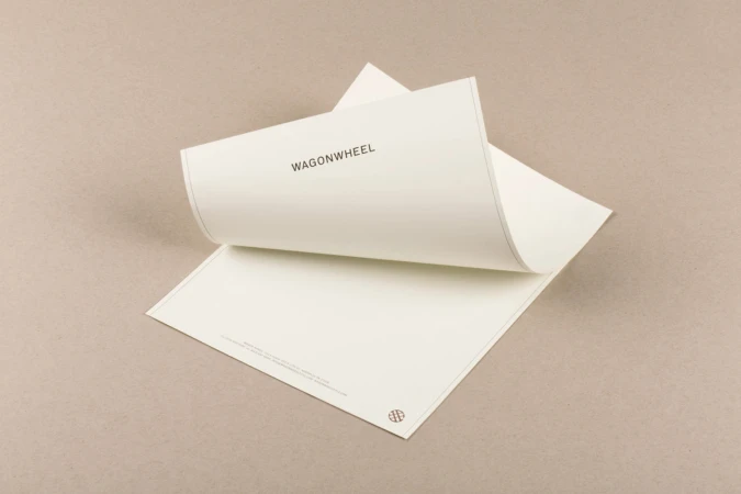 Image of two sheets of white paper with the text 'WAGONWHEEL' in a light neutral background. The top sheet is being lifted.