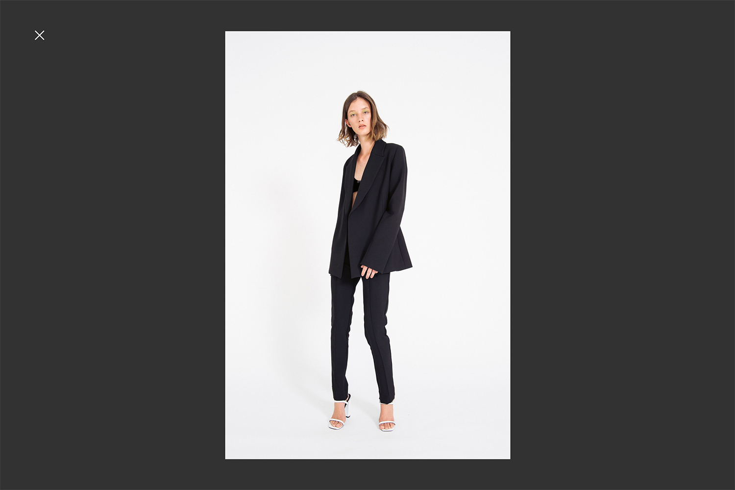 A fashionable young woman poses in a black blazer and pants, paired with white high heels, against a clean white backdrop.