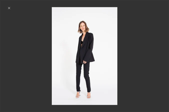 A fashionable young woman poses in a black blazer and pants, paired with white high heels, against a clean white backdrop.