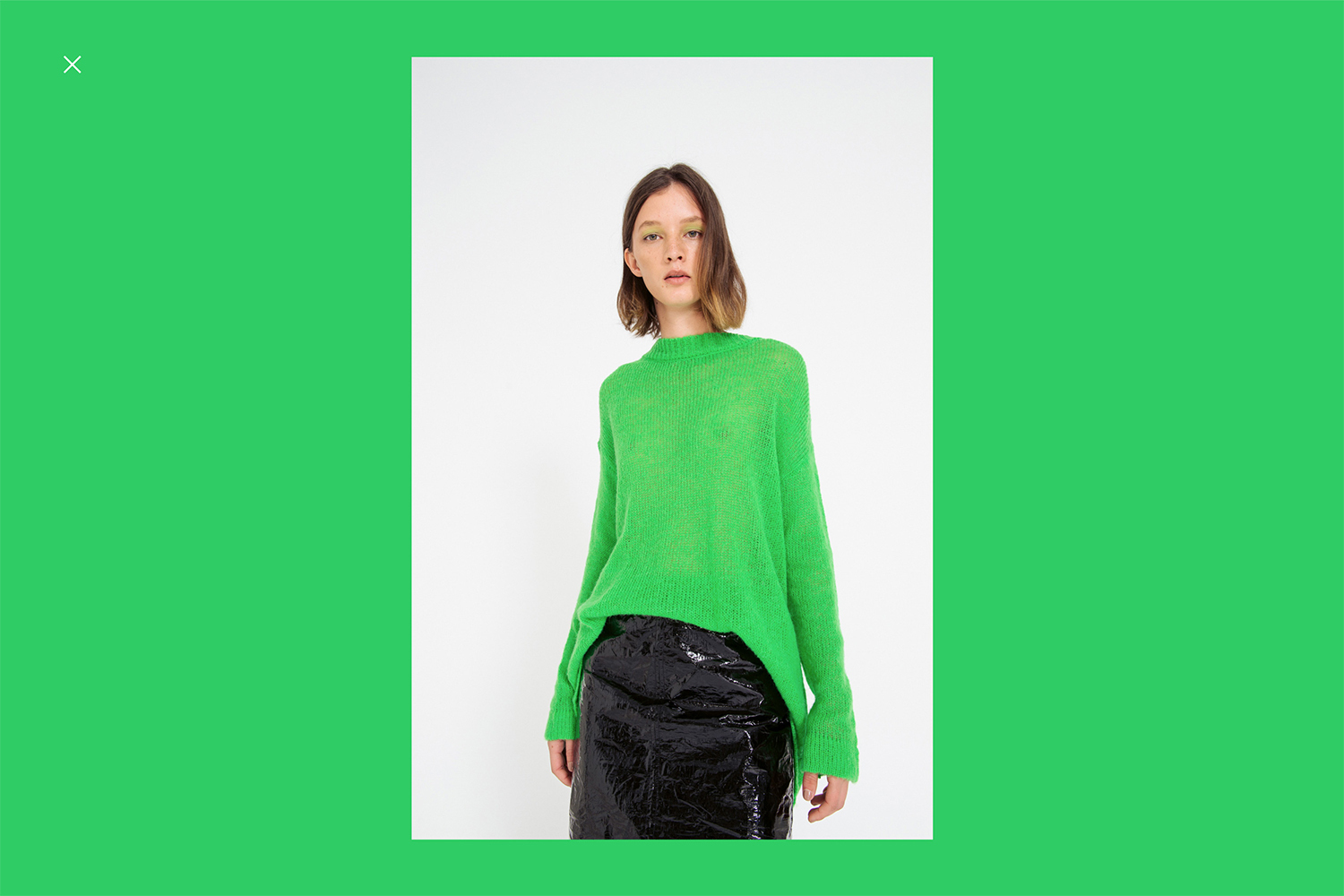 Woman wearing a green sweater and black skirt stands in a studio with bright lighting, showcasing fashion and style.