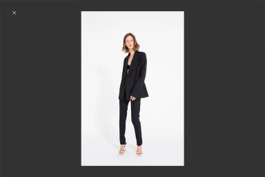 Model in a black suit and white heeled sandals standing against a plain white background for Georgia Alice