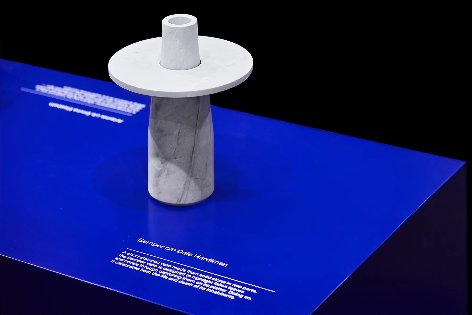 Sculptural stone vase art piece in two parts on display. The vessel is white, presented on a blue surface, with a black backdrop.
