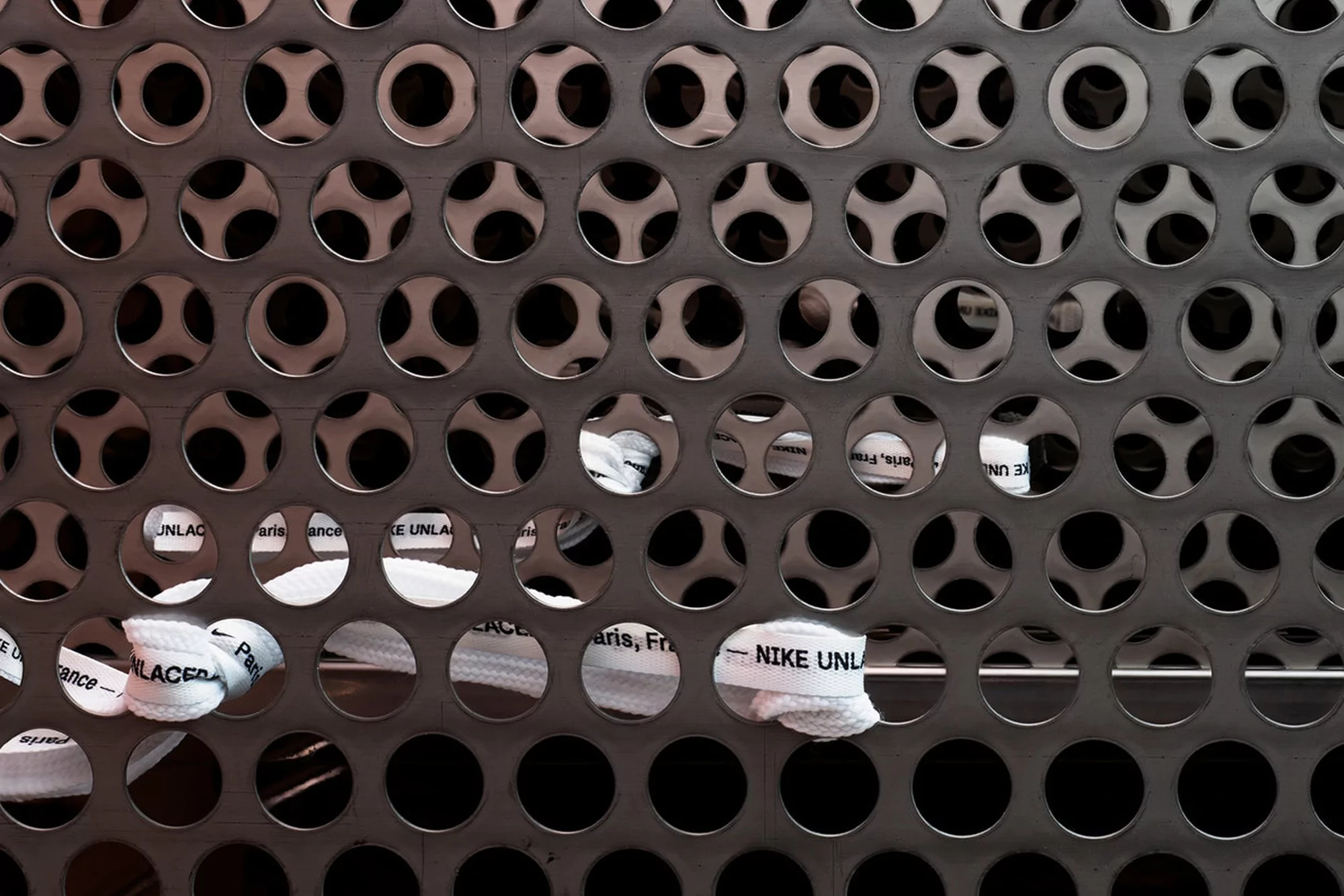 Close-up of patterned metal surface with sneaker laces visible through holes, featuring 'NIKE UNLACED' text, in a studio setting.
