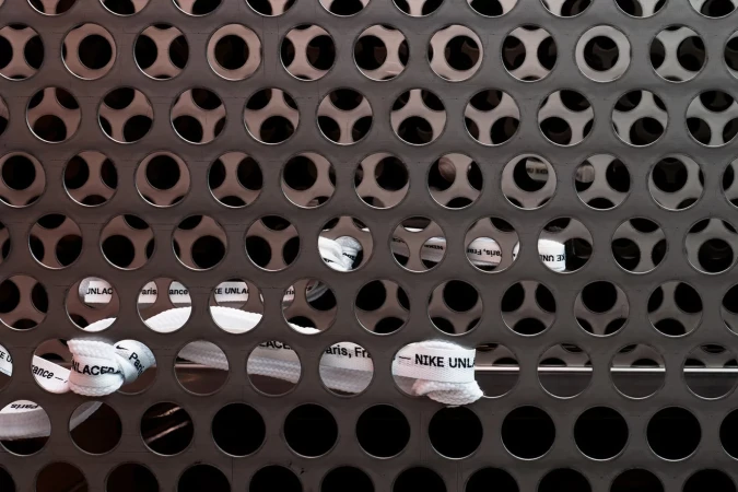 Close-up of patterned metal surface with sneaker laces visible through holes, featuring 'NIKE UNLACED' text, in a studio setting.