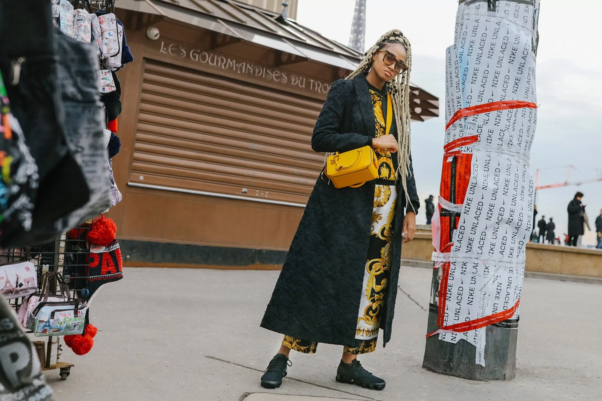 Stylish woman with yellow bag in Paris. She wears a long black coat over a patterned top and bottom, posing outdoors near the Eiffel Tower.