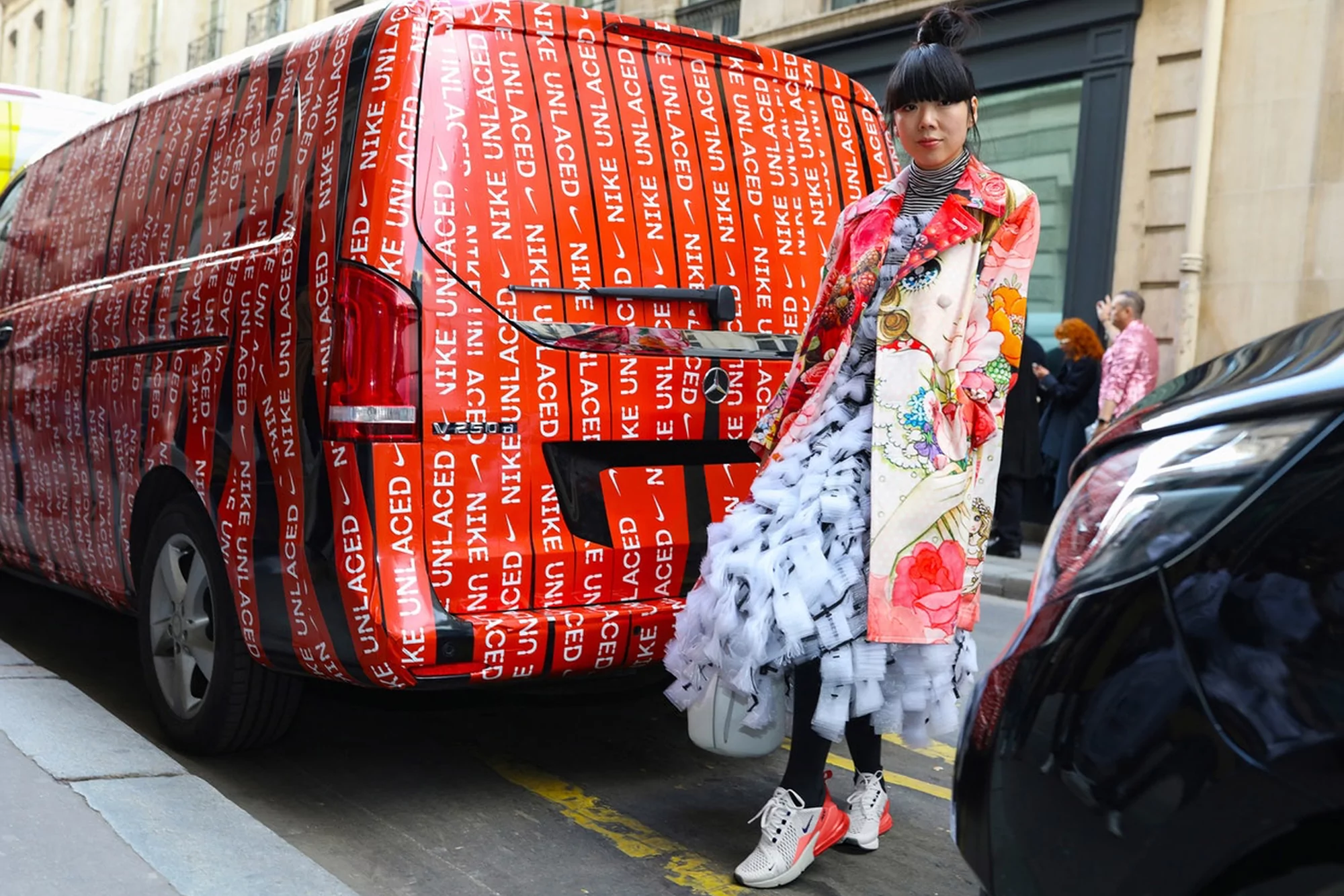 Asian woman in floral coat stands next to van covered in Nike Unlaced logos on a city street with a black car parked nearby.
