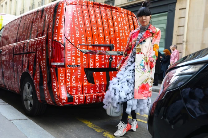 Asian woman in floral coat stands next to van covered in Nike Unlaced logos on a city street with a black car parked nearby.