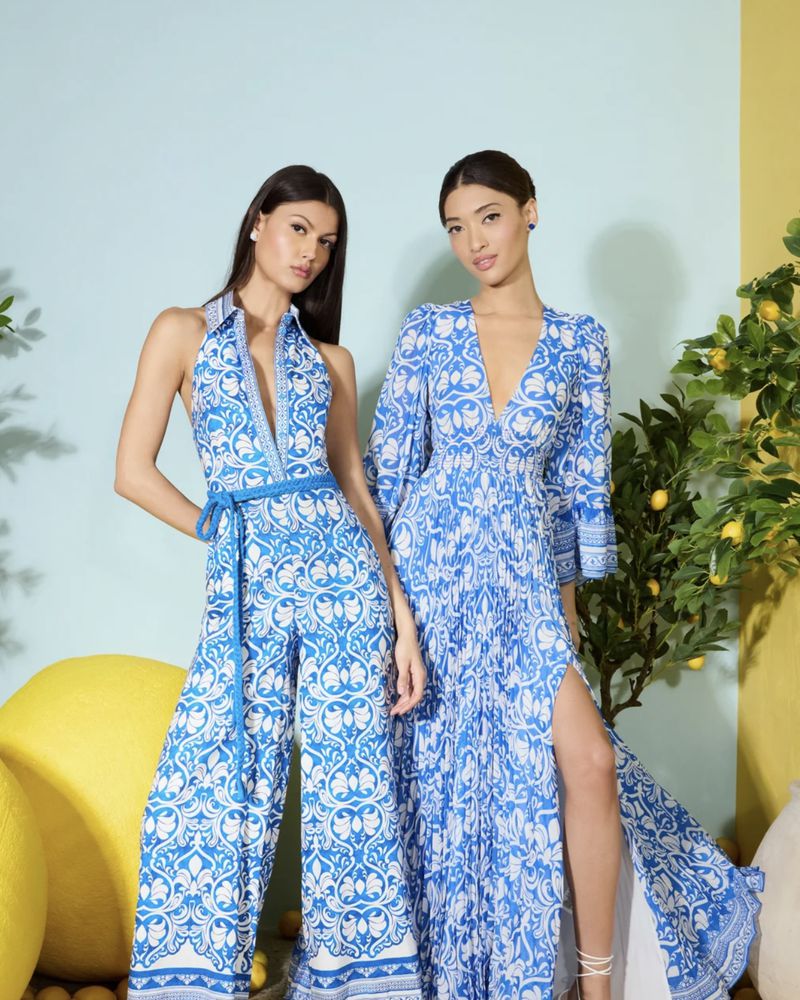 Two women pose in matching blue-and-white patterned jumpsuits and dresses against a studio backdrop with faux lemon tree accents.