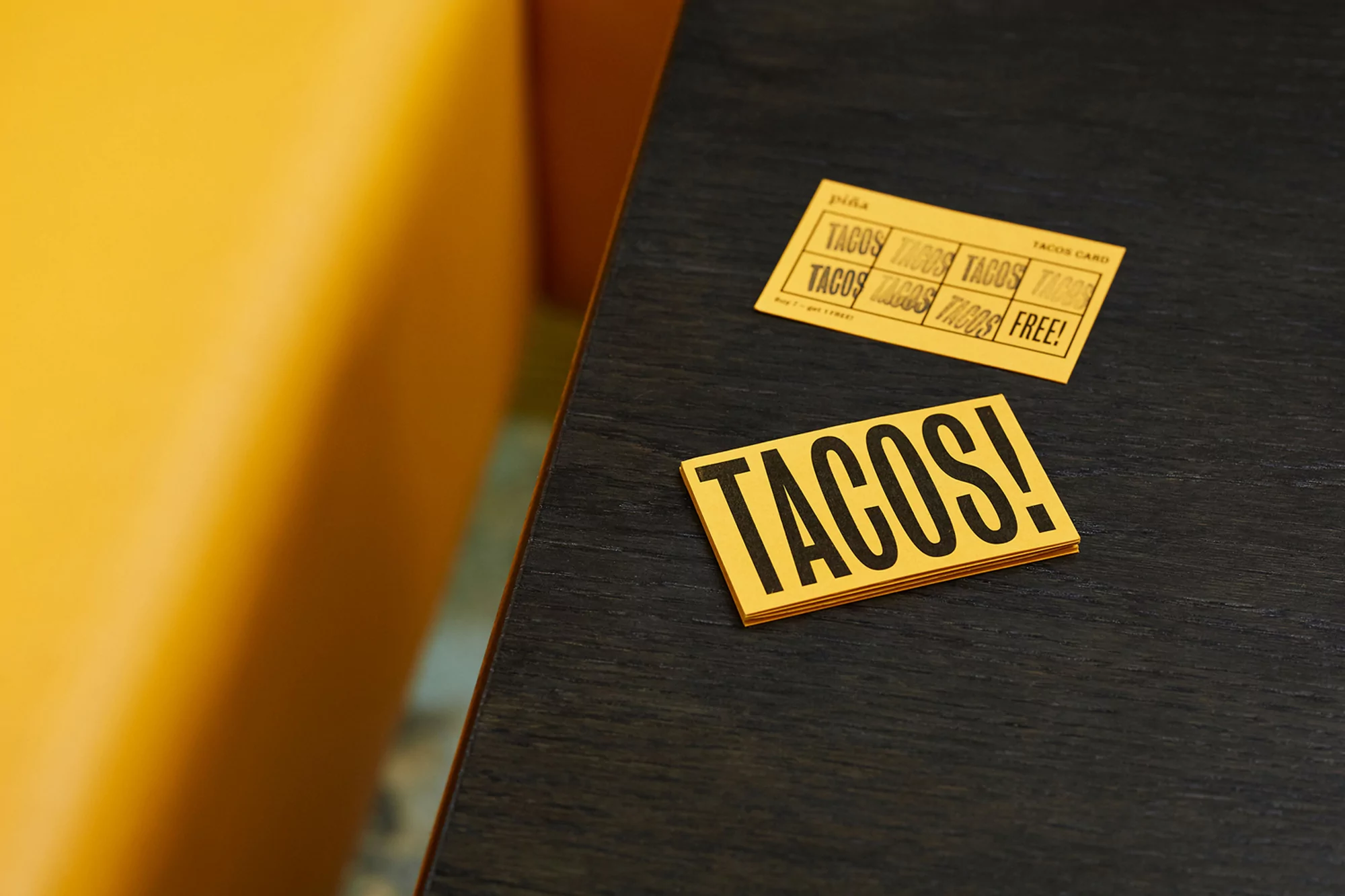 Close-up of taco cards on a dark wood table next to a yellow padded booth seat. The yellow cards have black text saying "TACOS!"