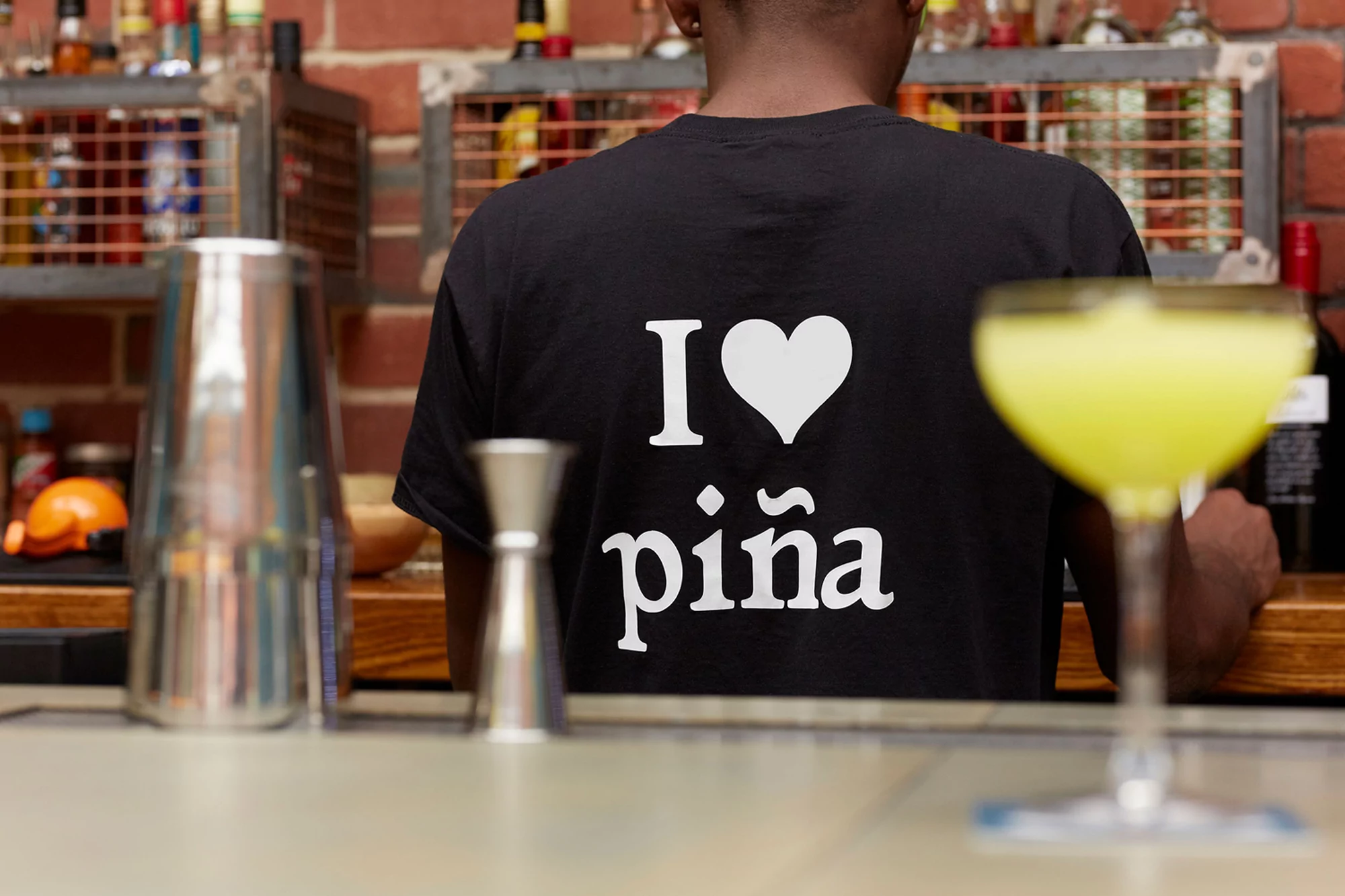 Bartender in black t-shirt with 'I <3 piña' stands behind bar with cocktail shaker and drink. Bar shelves and brick wall in background.