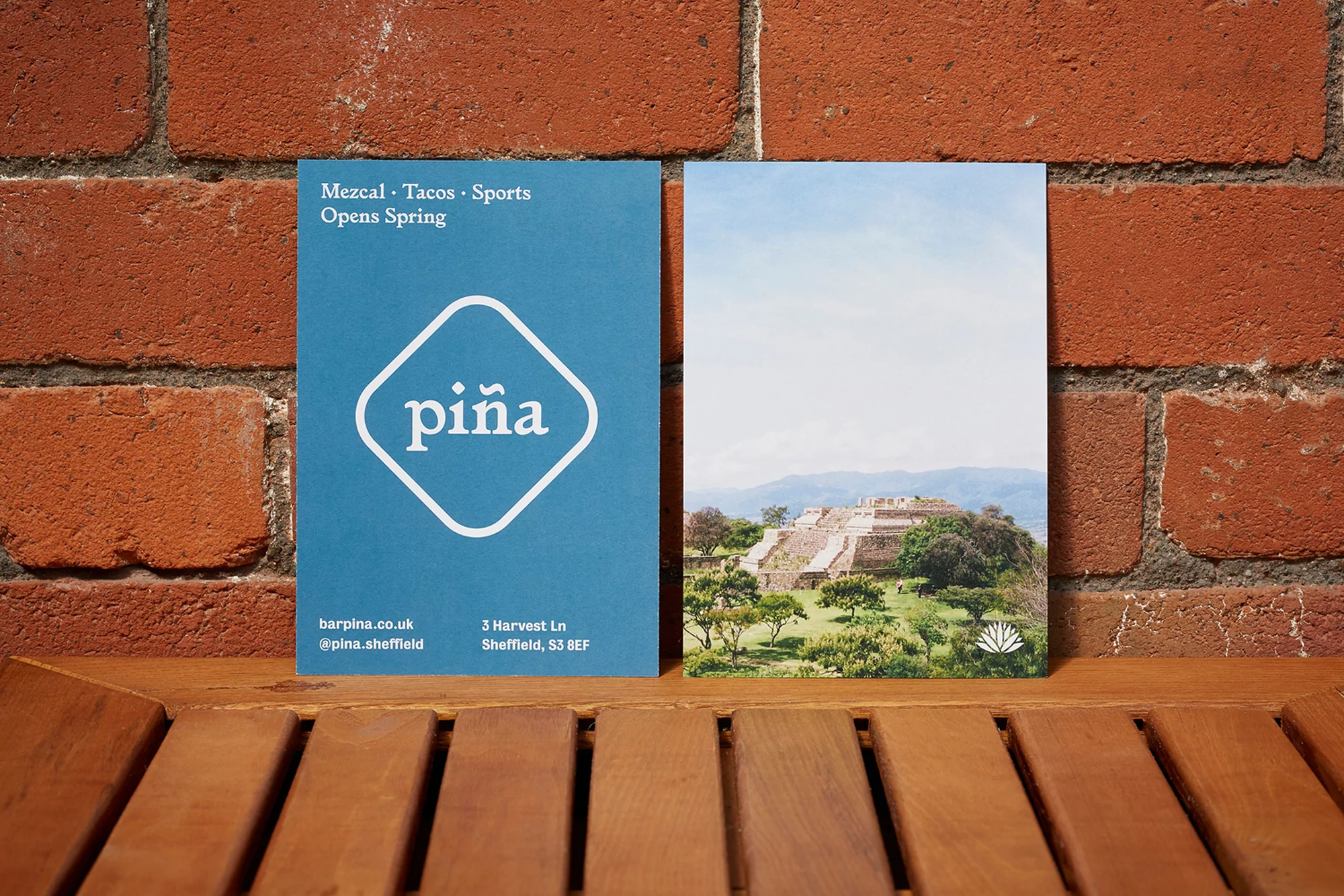 Two postcards on a wooden table against a brick wall. One is teal with the word 'piña' on it, and the other has a landscape.