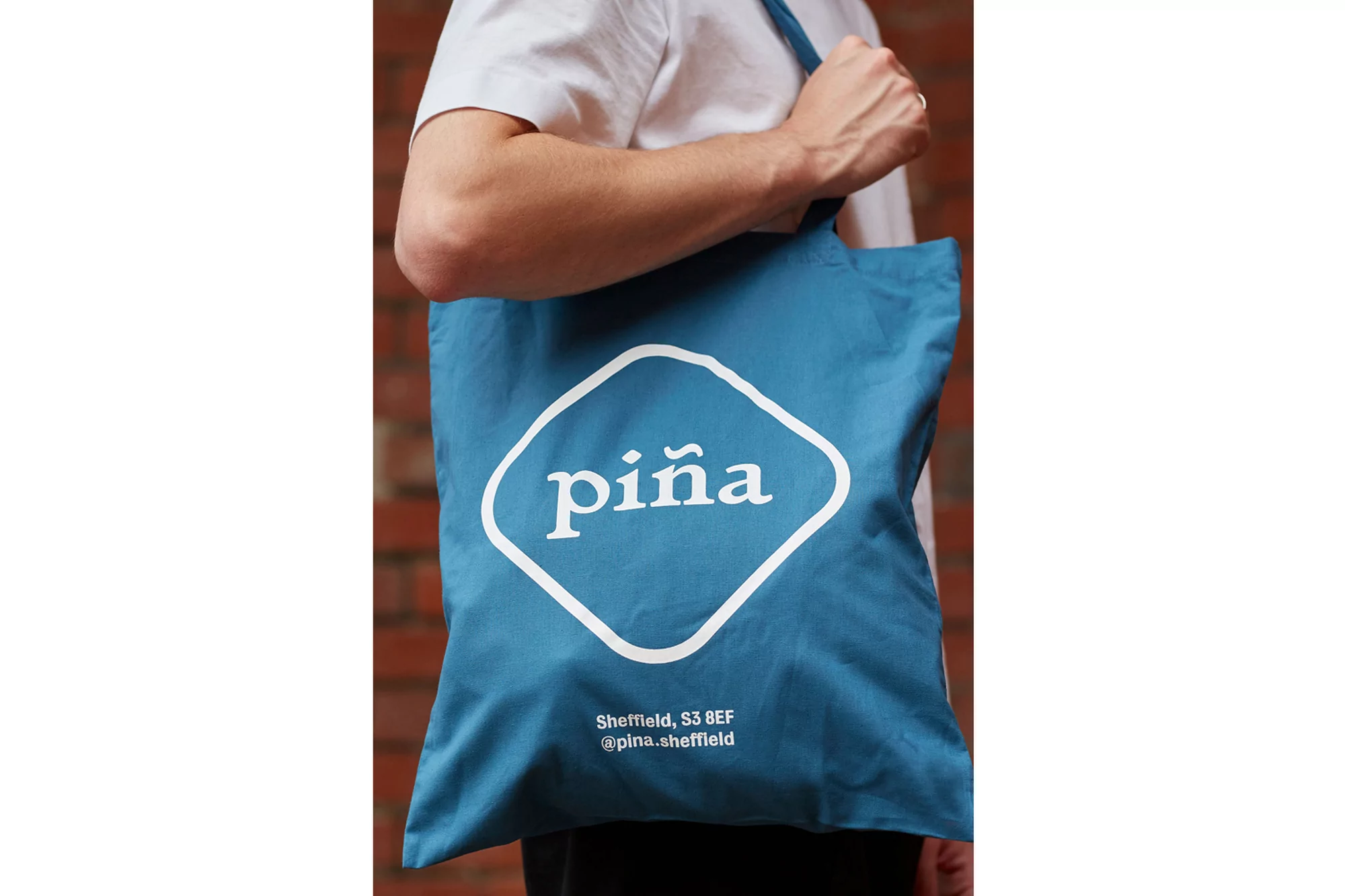Person holds a turquoise tote bag with the white 'piña' logo on a brick wall background, showcasing a brand and sustainable fashion.