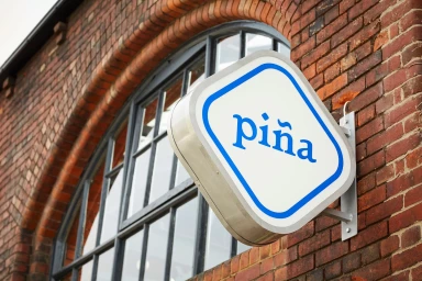 Square illuminated store sign with blue Piña logo mounted on a weathered red brick wall next to an arched window.