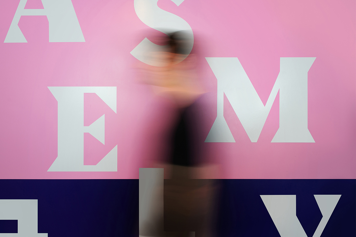 Motion blur of a person in front of a pink wall with white letters and a dark blue band at the bottom, creating an abstract effect.