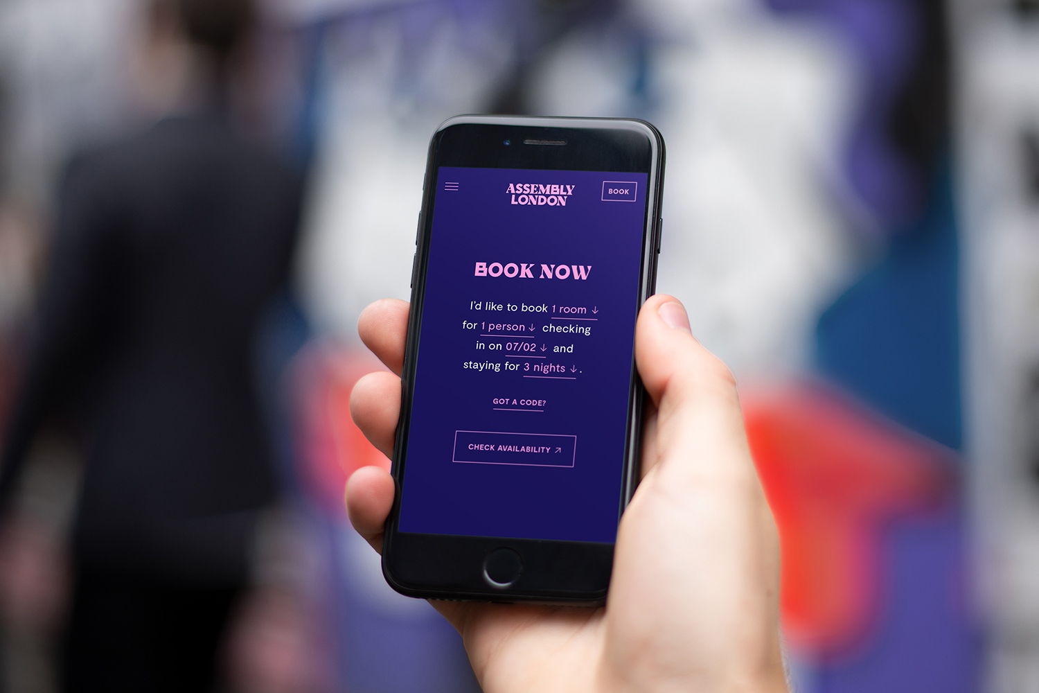 Person holds a smartphone displaying the booking screen for Assembly London hotel, featuring text and a purple background. Urban setting.