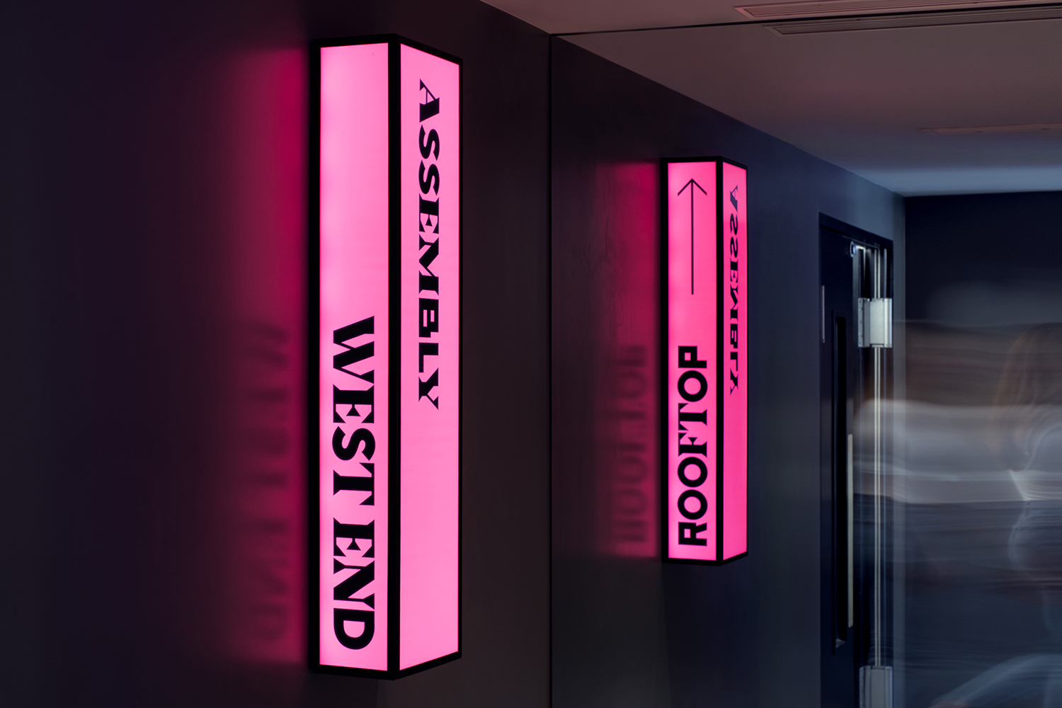 Illuminated directory signs on a wall point to 'Assembly' and 'Rooftop', offering directions in a modern, stylish indoor setting.