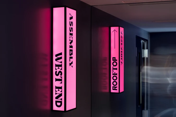 Illuminated directory signs on a wall point to 'Assembly' and 'Rooftop', offering directions in a modern, stylish indoor setting.