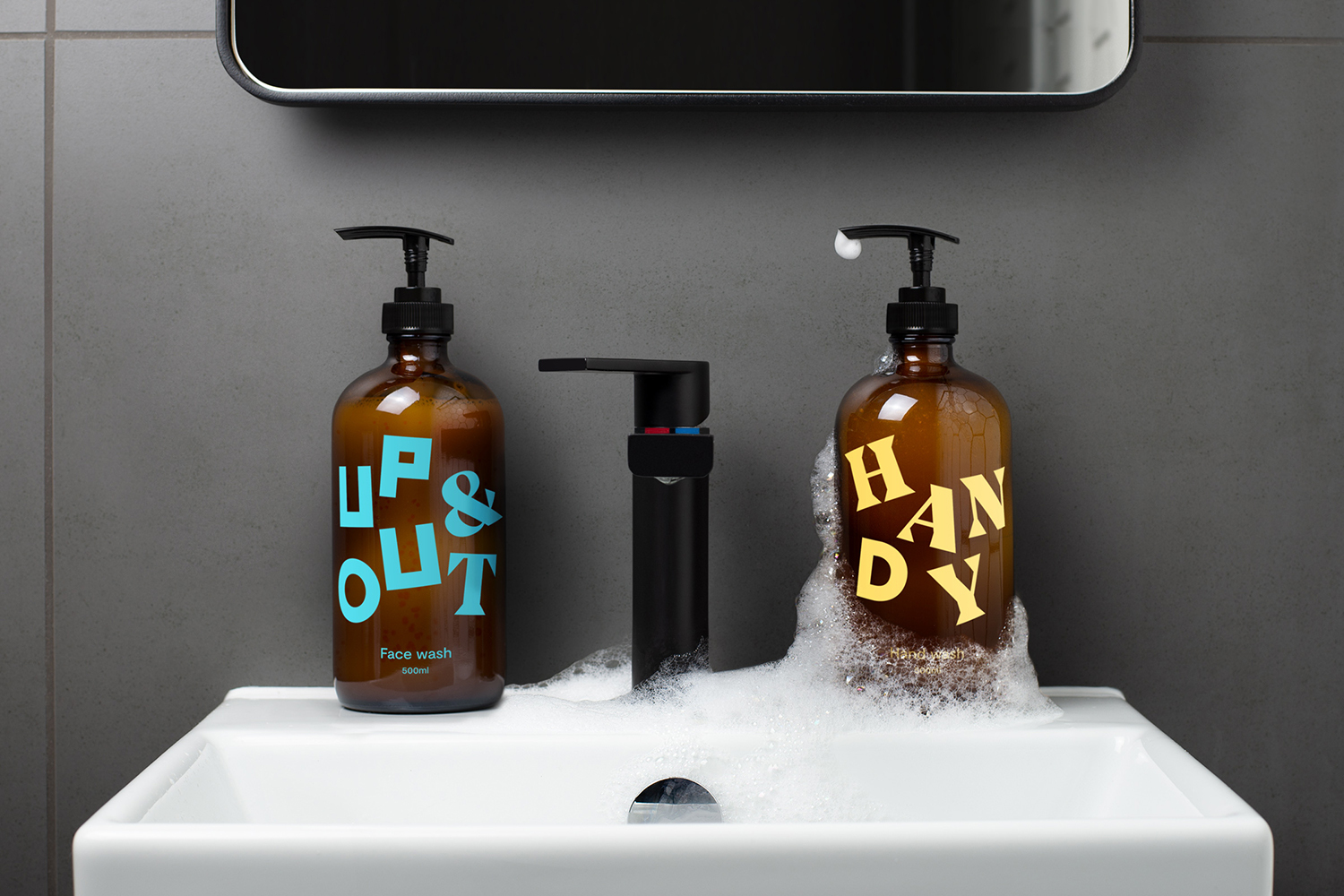 Two amber glass pump bottles with branding next to a black faucet on a white sink, against a gray tiled wall and a dark gray mirror.