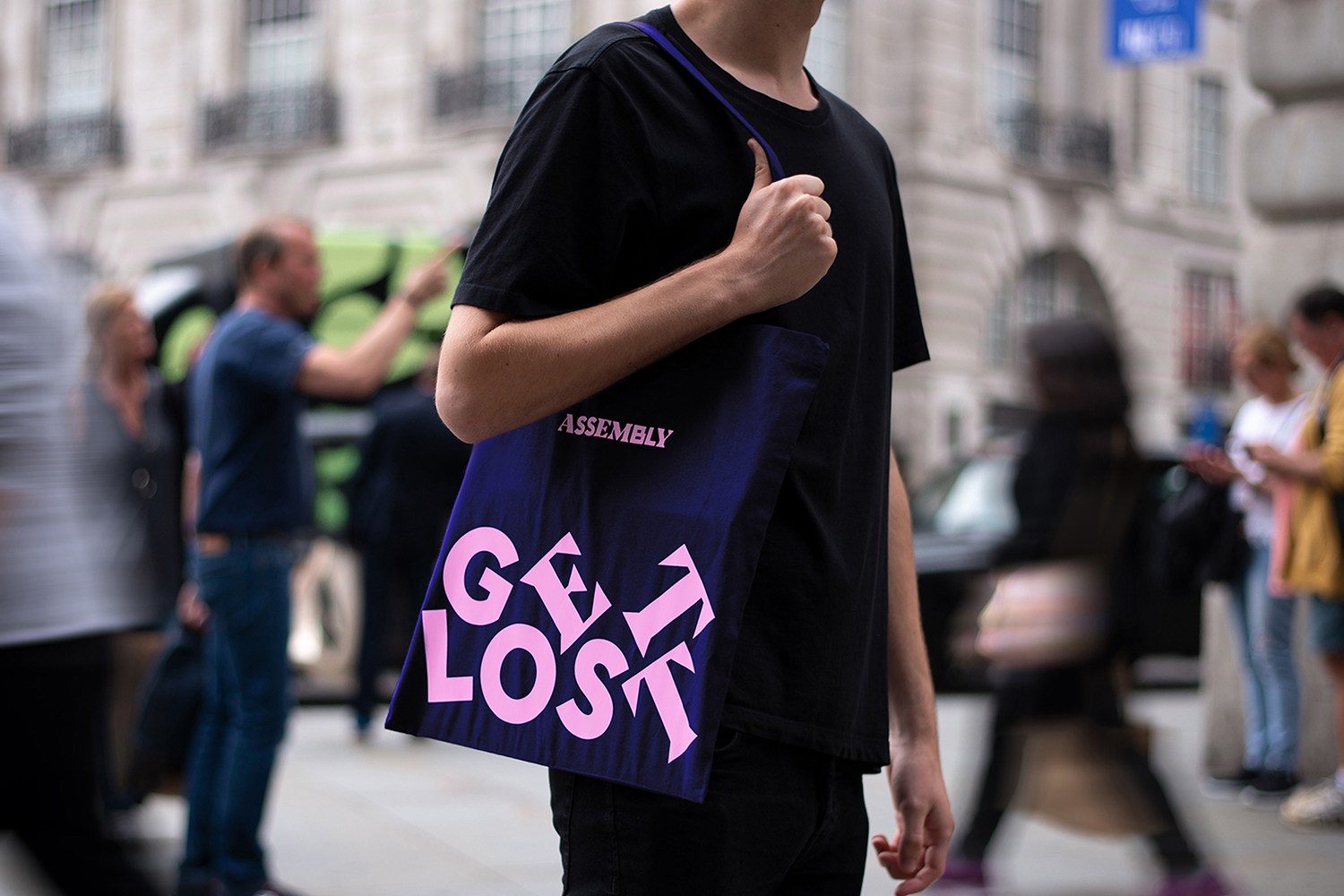 A person wearing a black t-shirt carries a blue tote bag with pink text 'GET LOST' in an urban outdoor setting with blurred pedestrians.