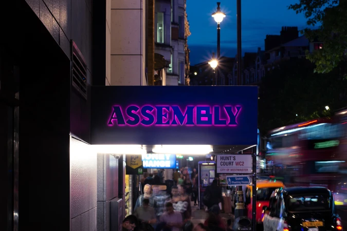 Urban scene at night features the Assembly building sign with a crowd of pedestrians and blurred motion of a bus and cars.