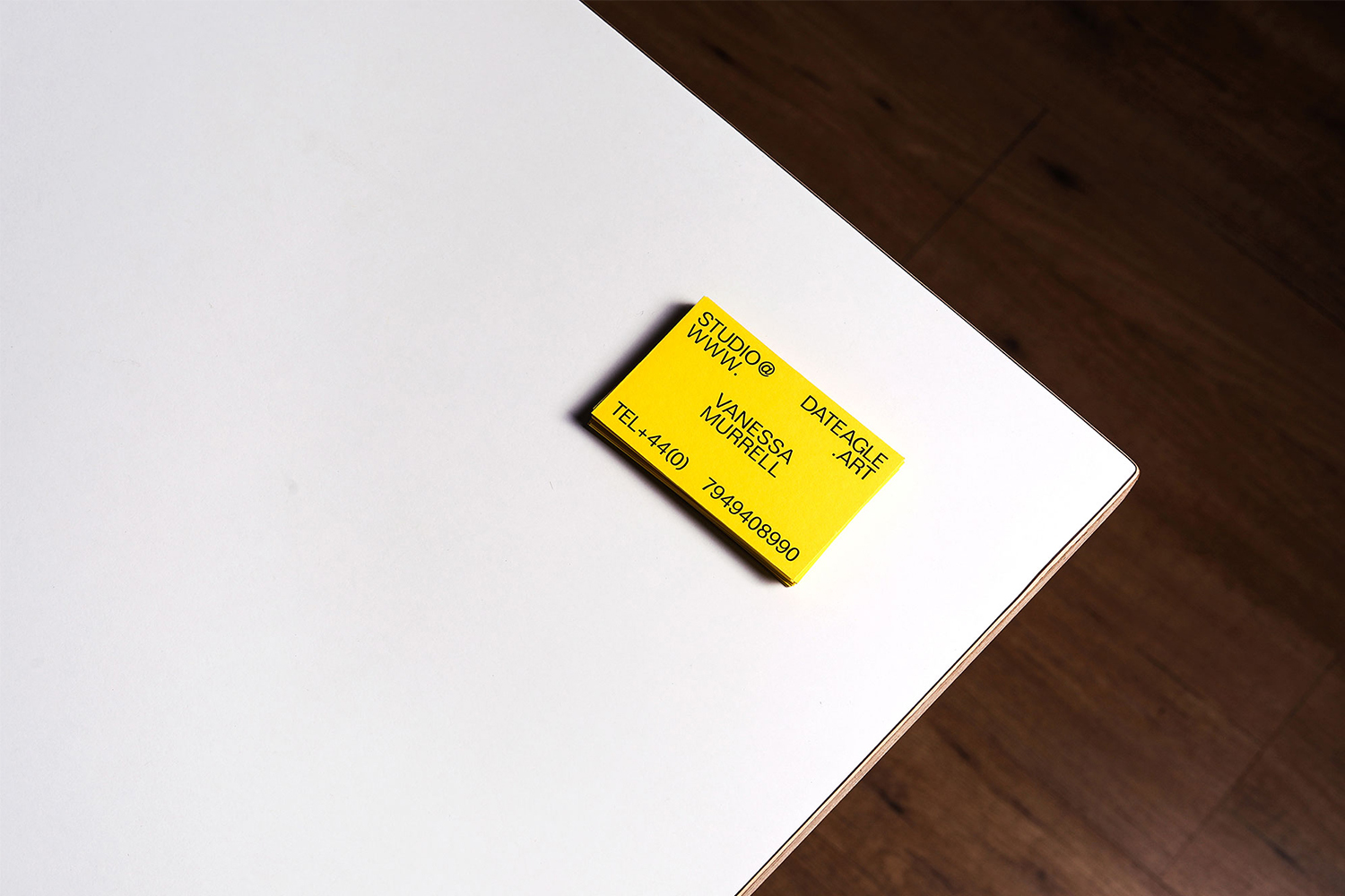 Bright yellow business card on a stark white table corner with dark wood floor background. Contact info, phone number visible.