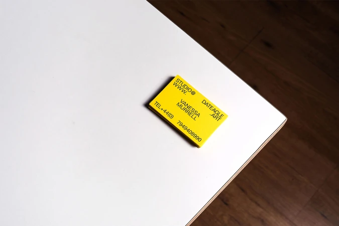 Bright yellow business card on a stark white table corner with dark wood floor background. Contact info, phone number visible.