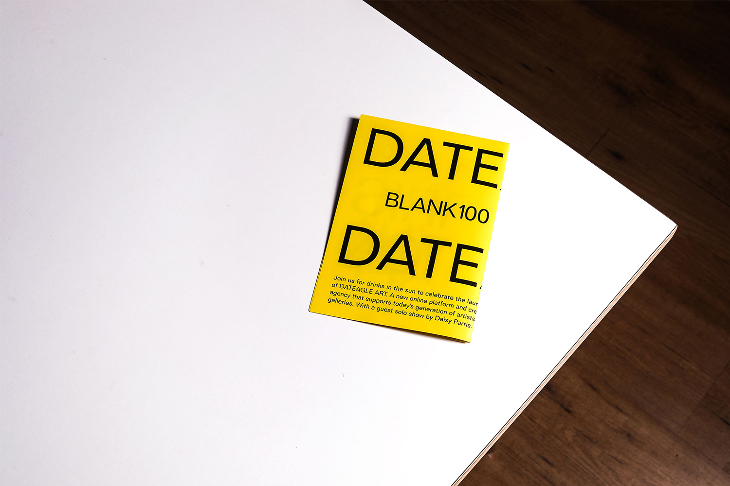 Close-up of a yellow flyer with the word 'DATE' printed in black, set on a white table with a dark wood floor backdrop.