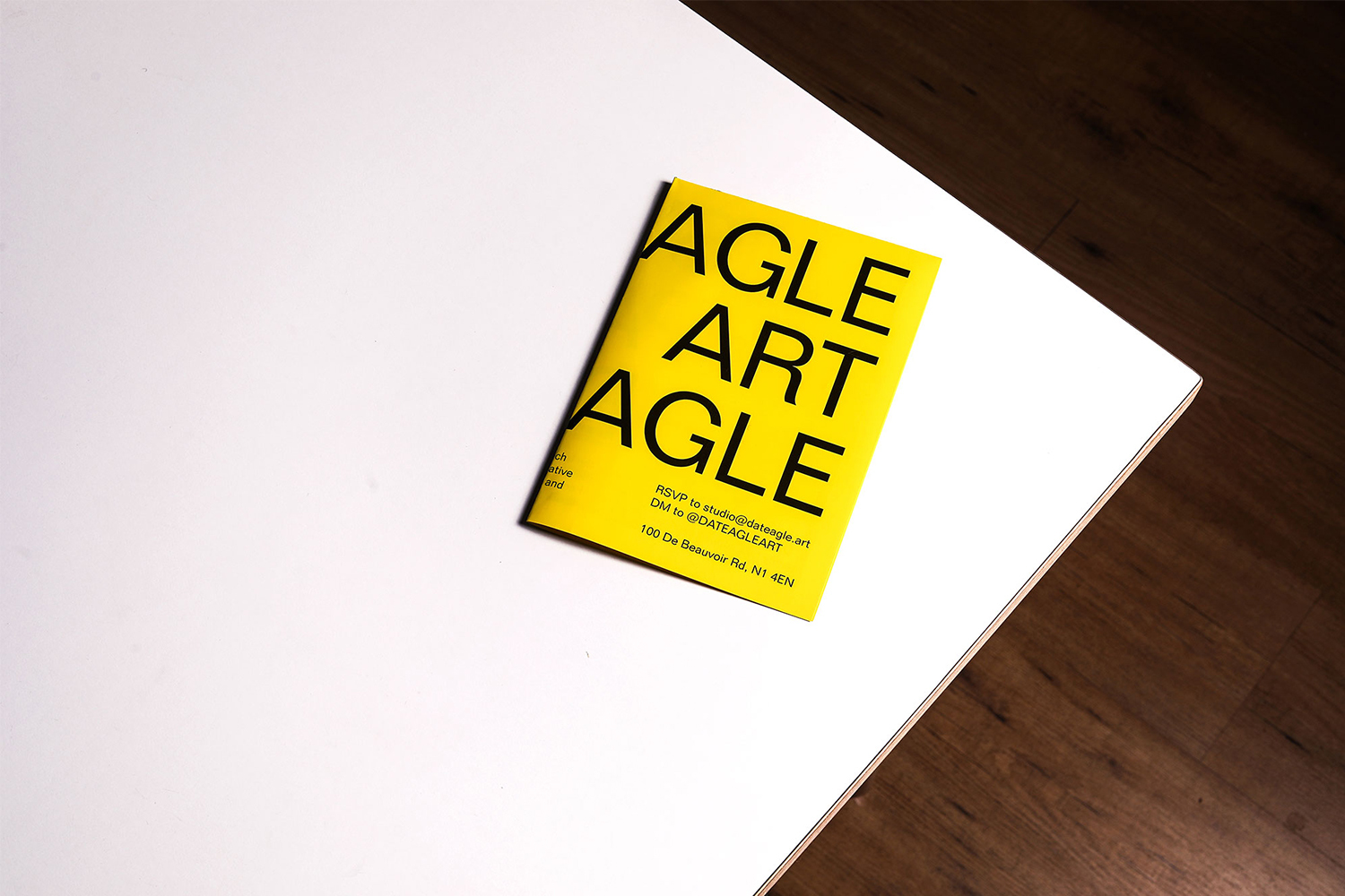 Promotional flyer with text 'AGLE ART AGLE' in bold yellow on a white tabletop near a wood floor, providing contact information.
