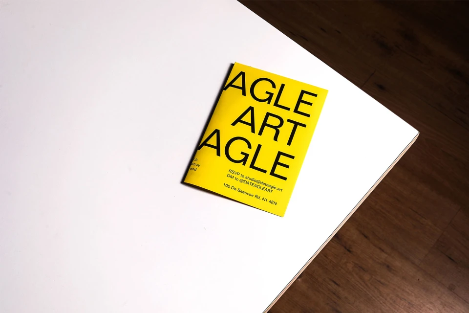 Promotional flyer with text 'AGLE ART AGLE' in bold yellow on a white tabletop near a wood floor, providing contact information.