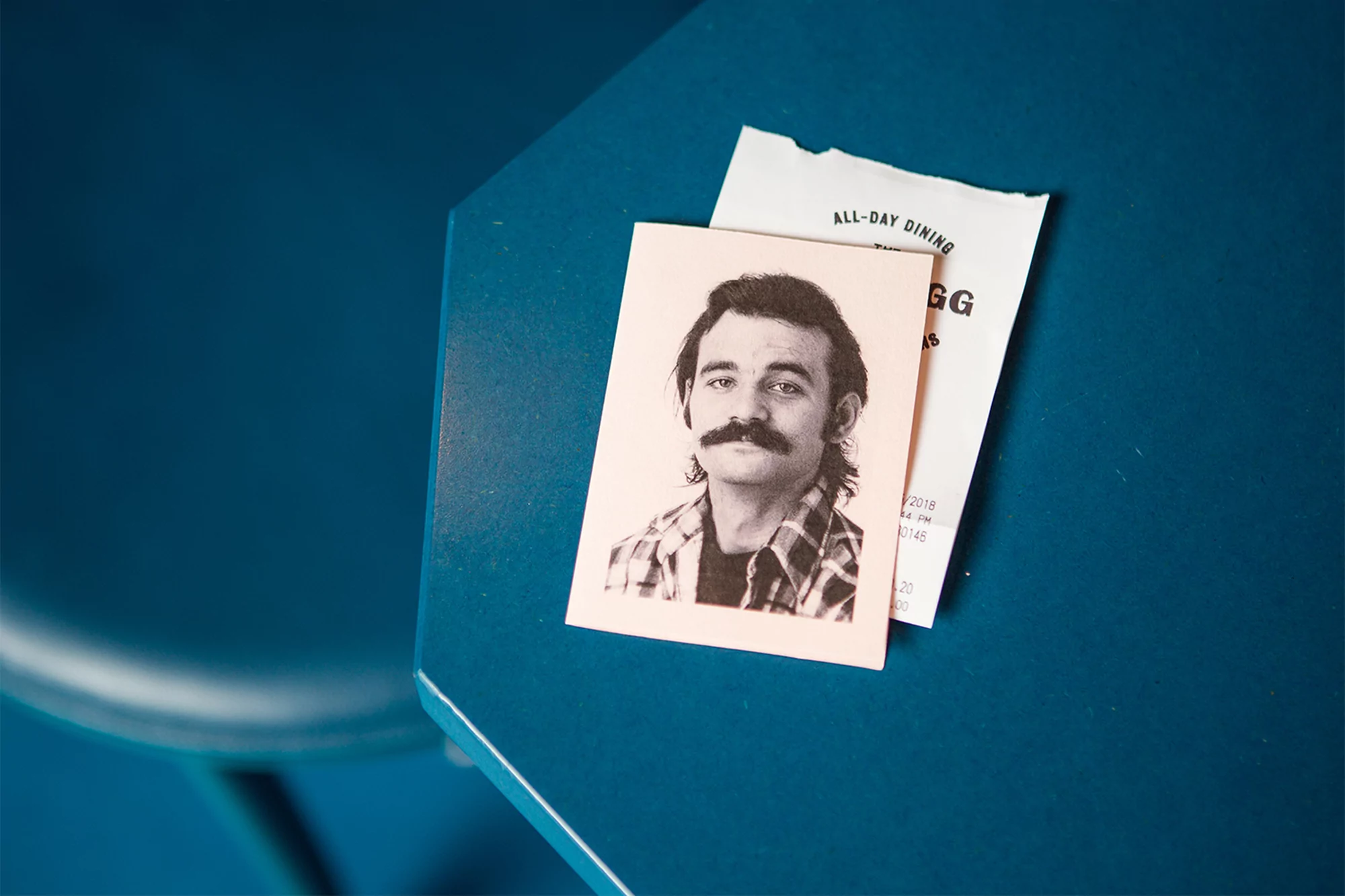 Photo of a man with a mustache on a card next to a receipt on a teal surface, showcasing portrait photography in a unique setting.