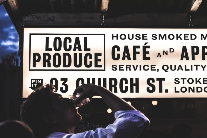 Close-up of person drinking a beverage below an illuminated cafe sign with local produce advertising and street address in London.