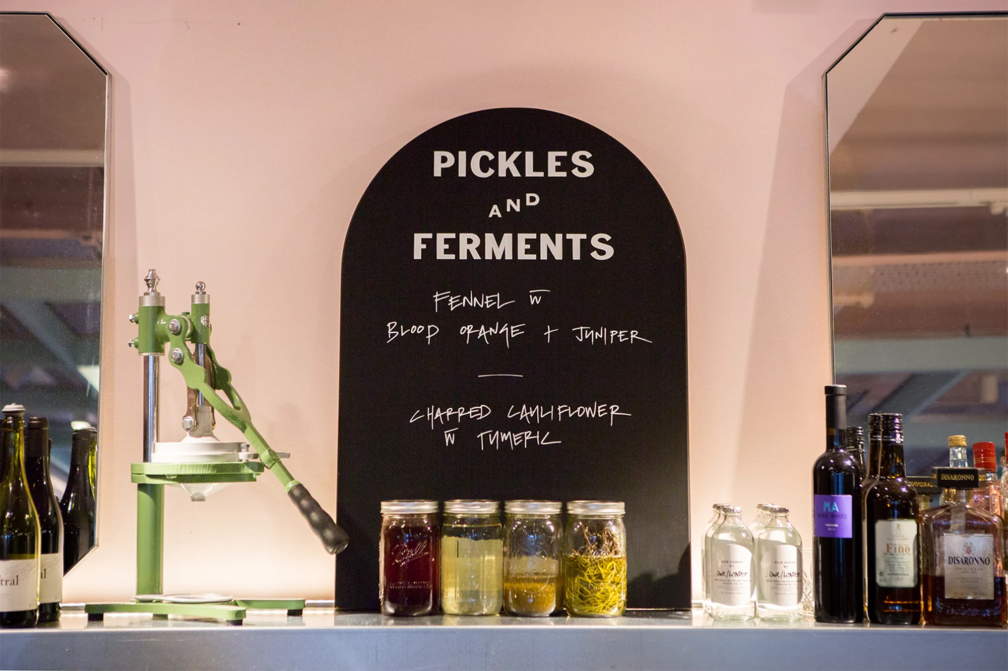 Close-up of a bar with wine and liquor bottles and a green food press near mason jars and a chalkboard sign with handwritten text.