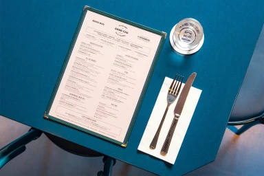 Overhead view of a restaurant menu, silver fork and knife on a white napkin, and a glass on a teal table