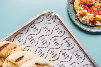 Overhead food photography of flatbread on branded parchment paper and a plate of whipped dip with toppings on a blue background