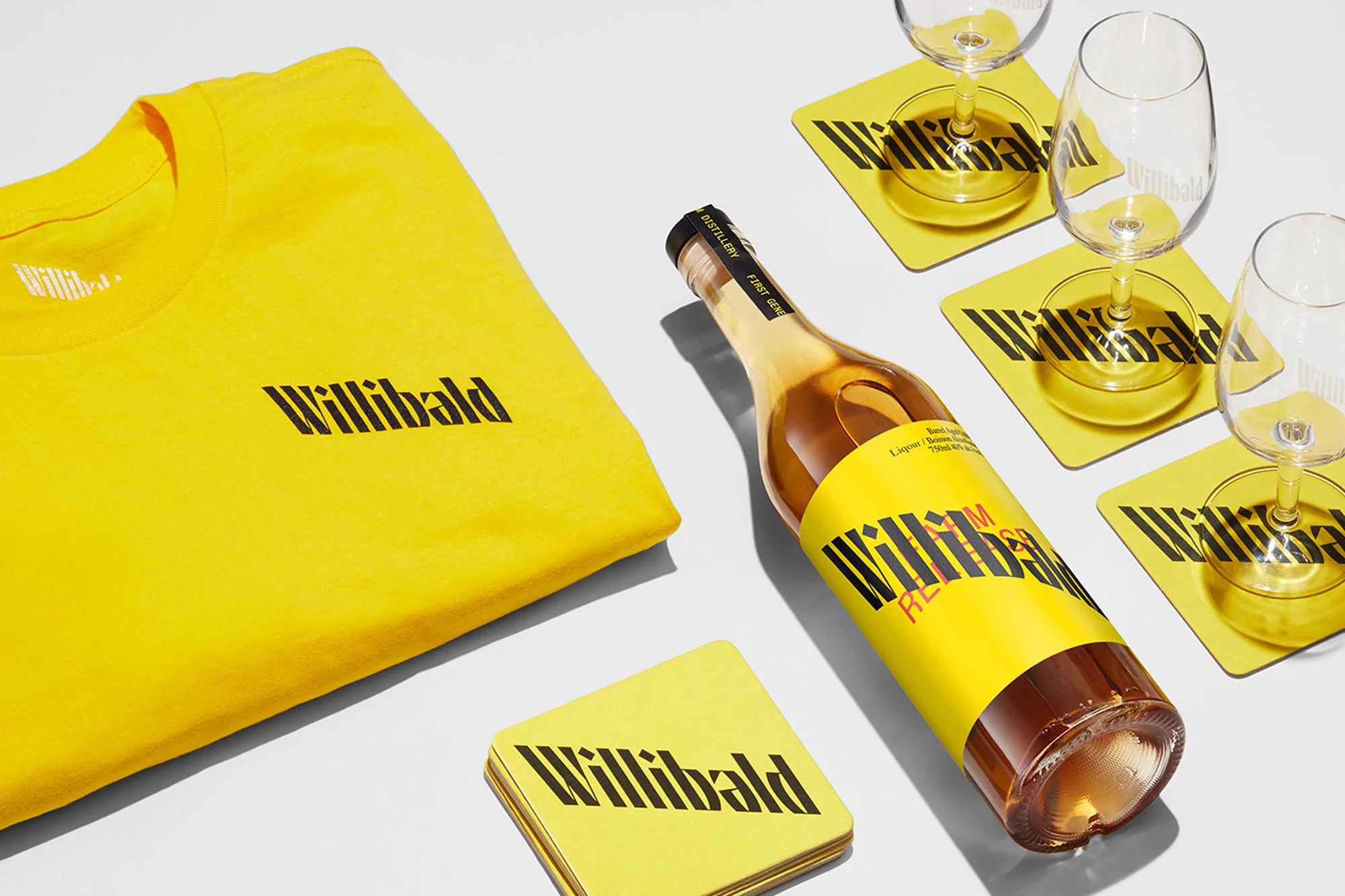 Still life with a bottle of Willibald gin, T-shirt, coasters, and glasses, all featuring the Willibald logo in black on yellow.