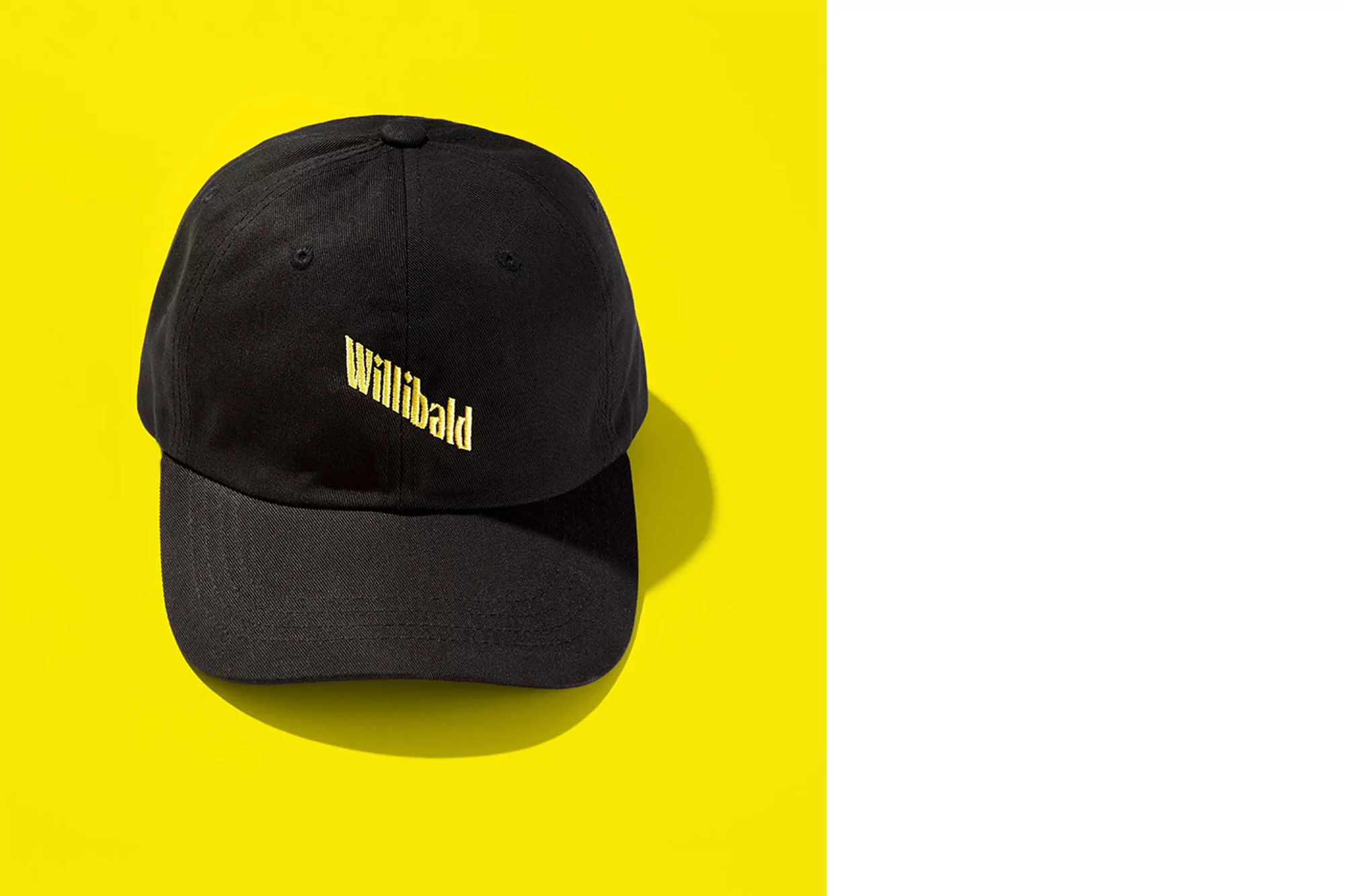 Black cap with 'Willibald' logo on yellow background. Product shot of a baseball cap. Minimalist aesthetic, clean lines, contrasting colors.