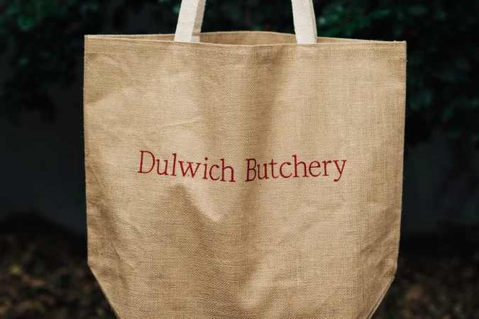 Jute tote bag with Dulwich Butchery logo against a blurred outdoor background showcasing sustainable and eco-friendly design.