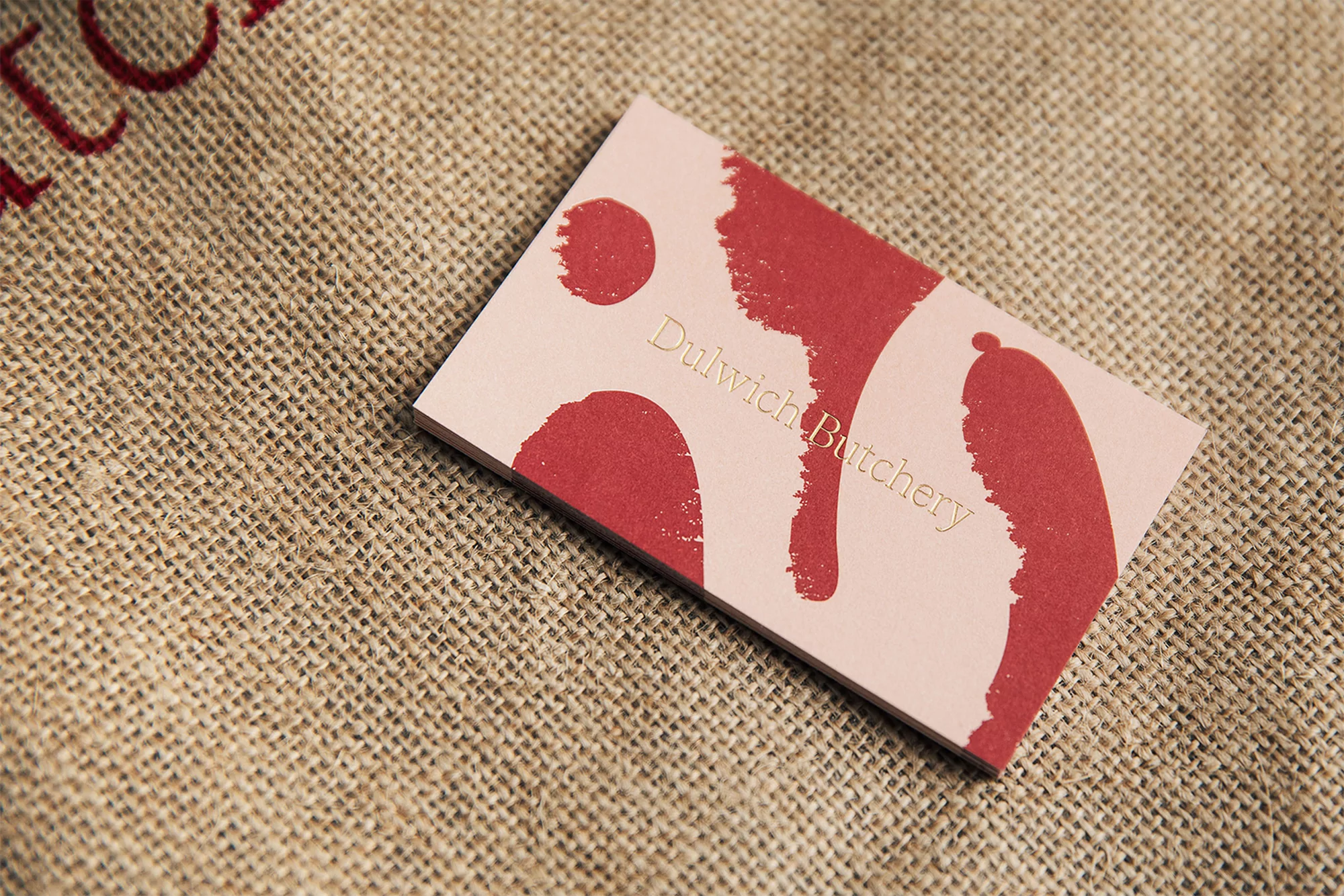 Close-up of a pink business card for Dulwich Butchery with organic red shapes, set against a textured burlap sack background.